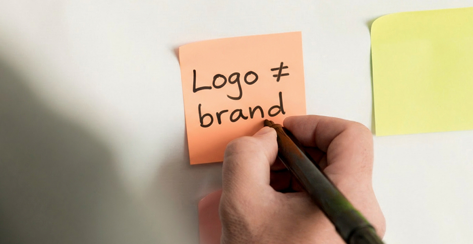 Eine Person schreibt mit einem Stift auf ein orangefarbenes Haftnotizzettel die Worte „Logo ≠ brand“.