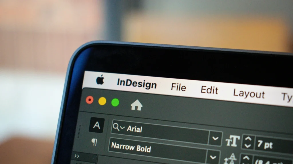 Nahaufnahme eines Bildschirms mit geöffneter Adobe-InDesign-Oberfläche. Sichtbar sind der InDesign-Schriftzug in der Menüleiste sowie geöffnete Typografie- und Bedienfelder.